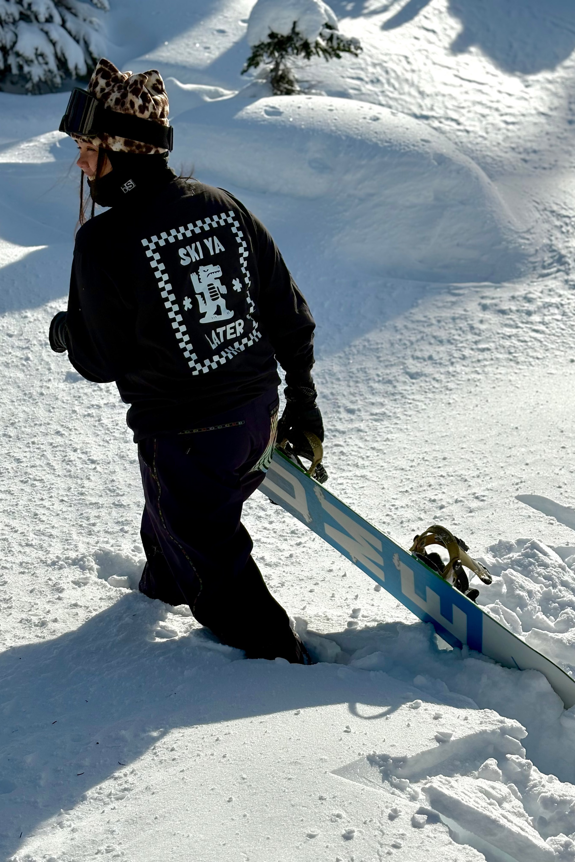 Person wearing a black jacket with a logo, leopard print beanie, and goggles on a snowy background
